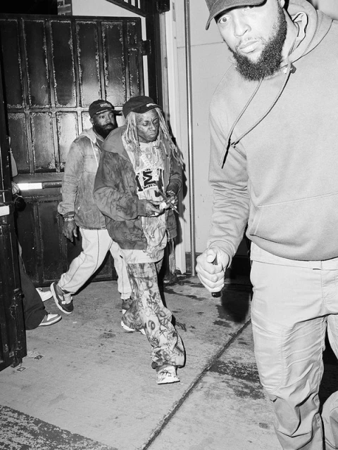 Black and white medium shot of Lil Wayne and his entourage walking out of a venue toward the camera in streetwear fashion.