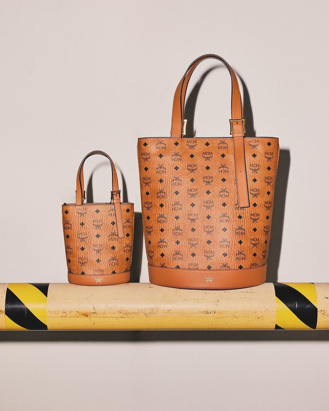 Two brown leather MCM bucket bags in small and medium size with all-over monogram print, sitting on a yellow and black industrial bar.