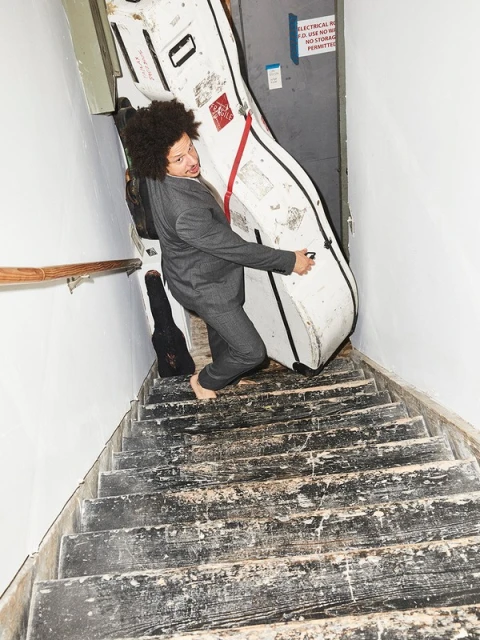 Man in suit carries oversized white musical instrument case down distressed stairs inside. Interior shot with natural light.