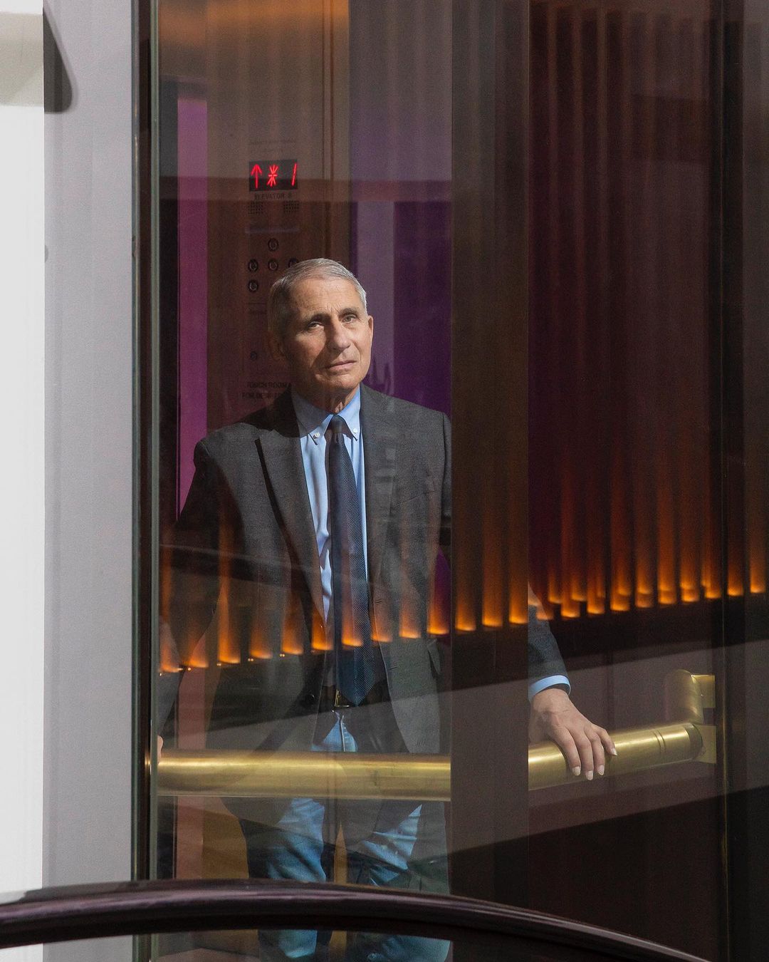 Adult male with gray hair and a suit stands in a glass elevator wearing jeans and a tie in front of a dark reflective wall.