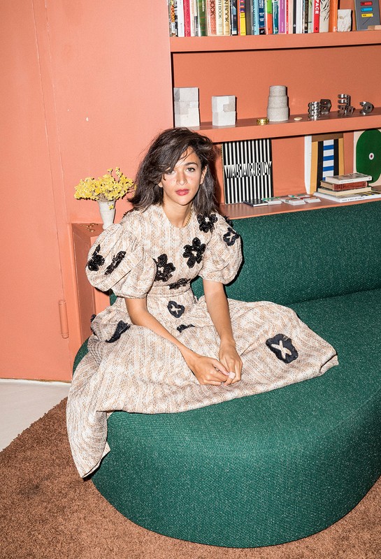 A woman with dark hair and a light complexion poses in a floral print dress on a green sofa in an indoor setting with books behind.