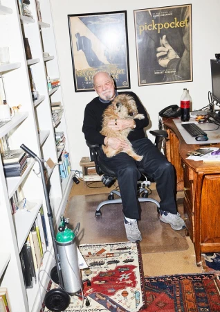 An older man with white hair and a beard is sitting in a chair, holding a small dog in an indoor office setting with posters on the wall.