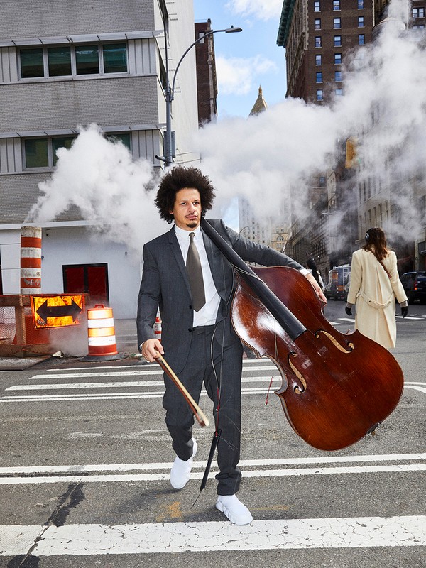 Eric Andre with a bass standing on a crosswalk in a city street with steam and buildings in the background on a bright day.