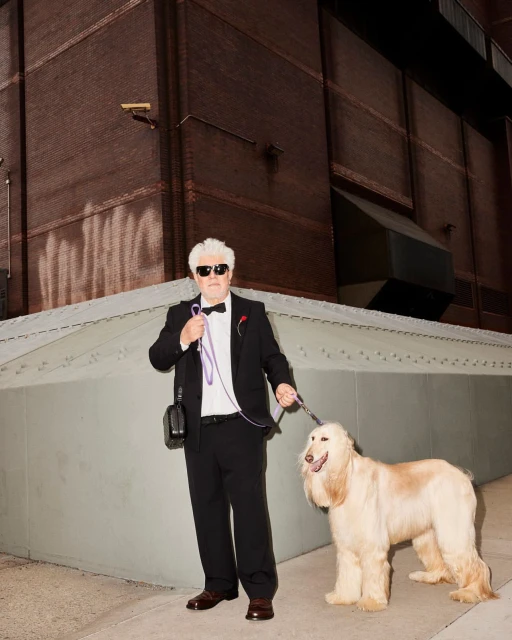 An adult male poses with an Afghan Hound dog on an urban street near a brick building, wearing a tuxedo, sunglasses, and holding a leash.