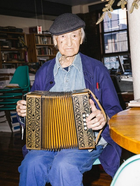 Elderly man in newsboy cap plays a Hohner accordion indoors with bookshelves and a table visible in the background.