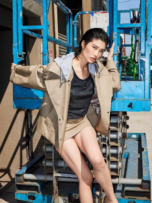 Adult female posing on blue scaffolding in outdoor urban setting wearing tan jacket, black top, tan miniskirt, tan heels.