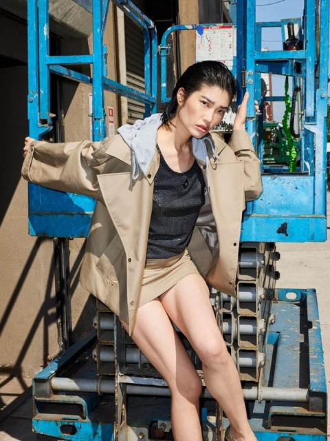 Adult female posing on blue scaffolding in outdoor urban setting wearing tan jacket, black top, tan miniskirt, tan heels.