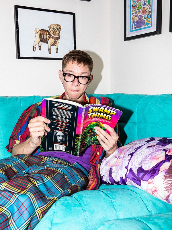 Young man with glasses reads a comic book titled Swamp Thing while sitting on a blue sofa in a room with framed wall art.