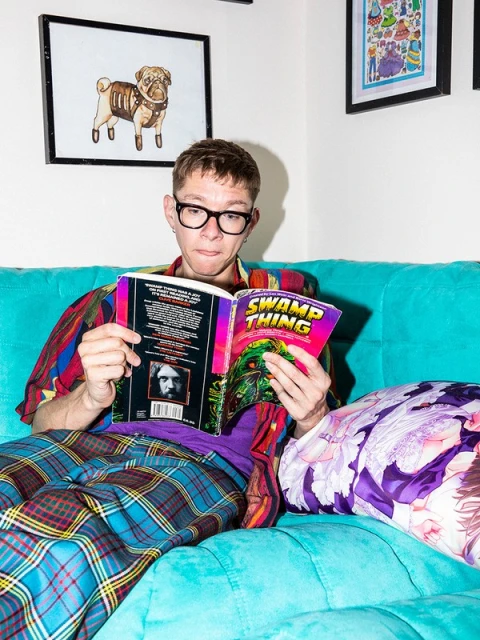 Young man with glasses reads a comic book titled Swamp Thing while sitting on a blue sofa in a room with framed wall art.