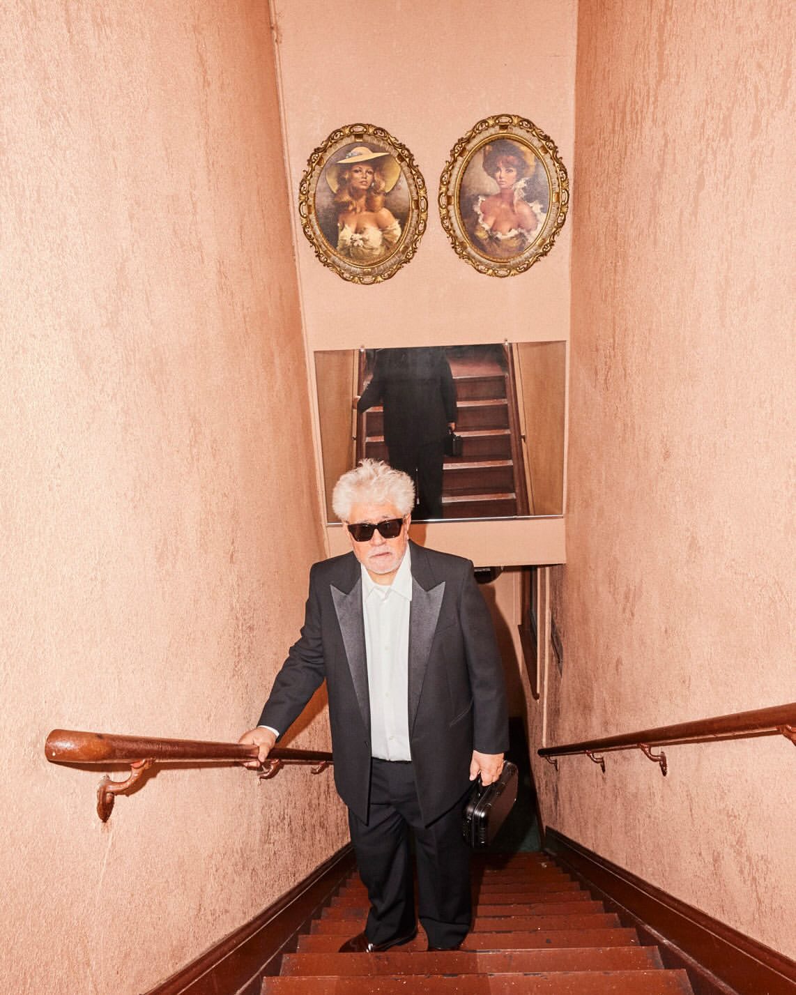 Adult male wearing a black tuxedo descending red stairs with a black briefcase indoors with a mirror and framed art on the pale-pink wall.