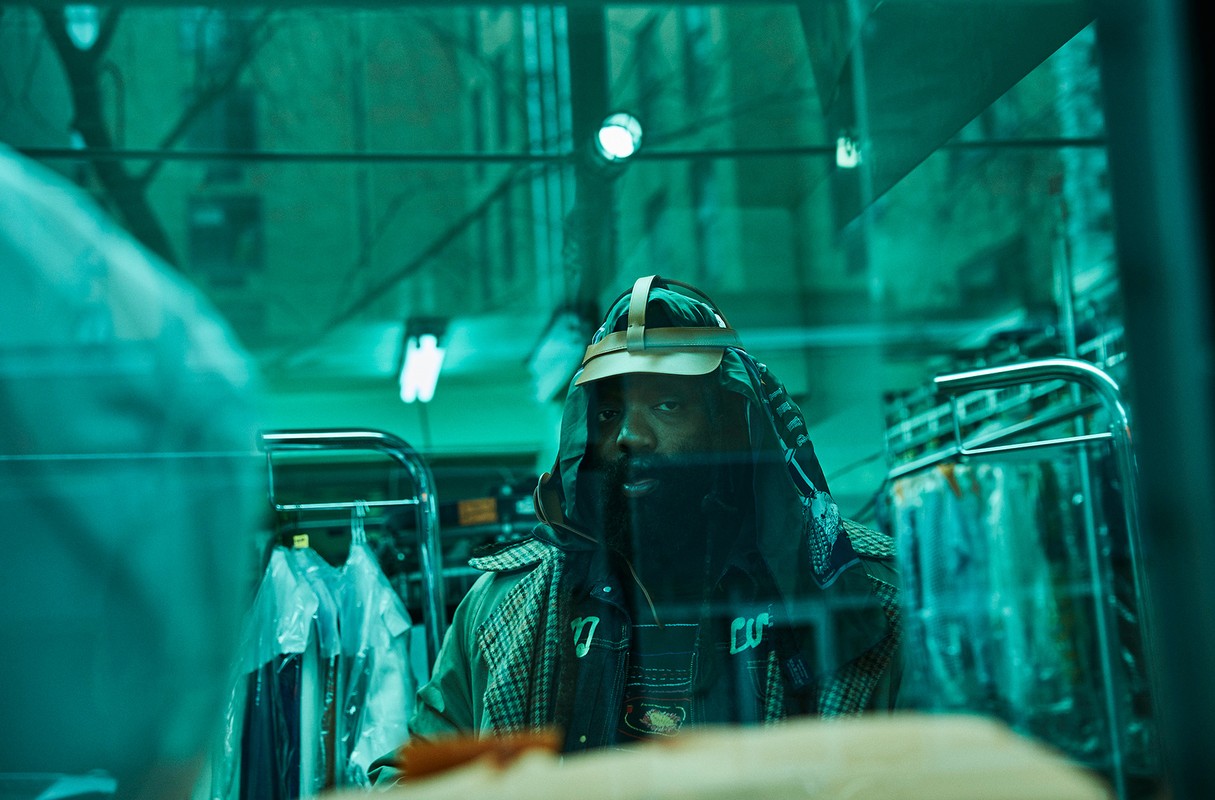 Adult male with a long beard wearing an embellished jacket and head covering standing in an indoor location that is teal coloured.