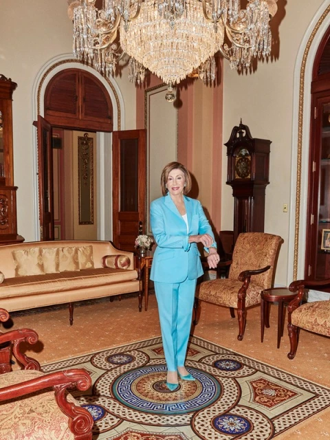 Full-length shot of a woman in a light blue pantsuit standing on an ornate carpet, with a large chandelier in the background.