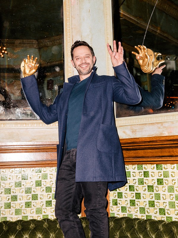Smiling man in navy coat posing with gold hand sculptures inside an ornate interior with decorative mirrors and olive green seating.