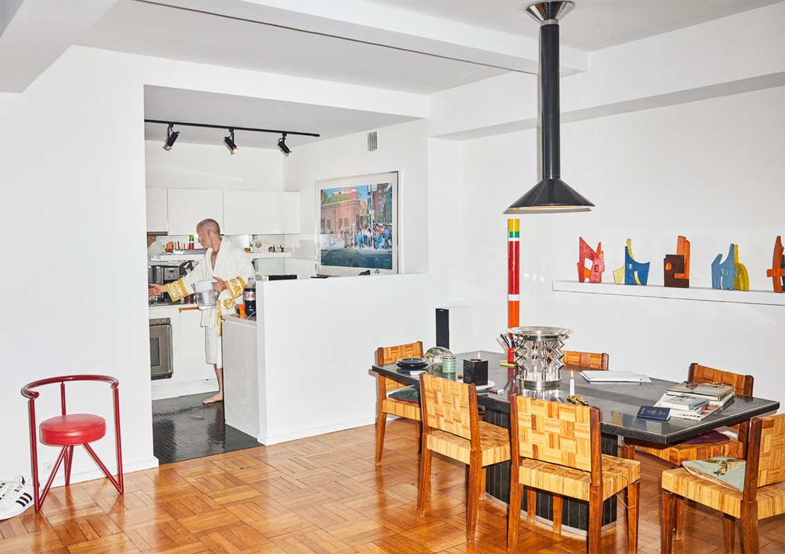 Adult male in a white Versace bathrobe cooks in a brightly lit, modern kitchen and dining area with wooden floors and black countertops.