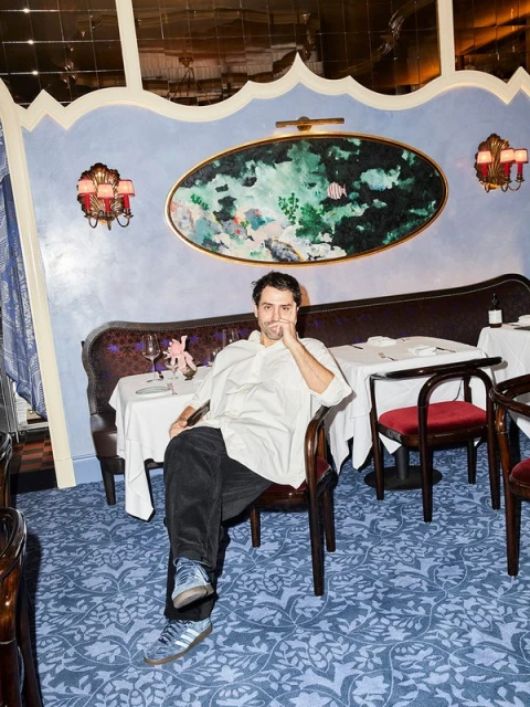 Man sitting in ornate restaurant, wearing a white shirt and black pants. The space features patterned carpet and elegant table settings.