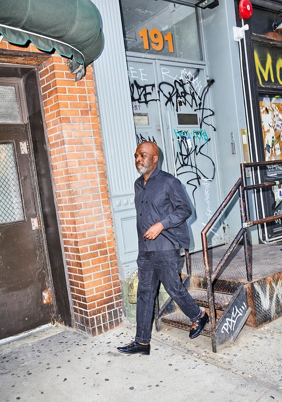 An adult male with a bald head walks down steps outside a brick building in an urban setting, wearing a dark shirt, jeans, and shoes.