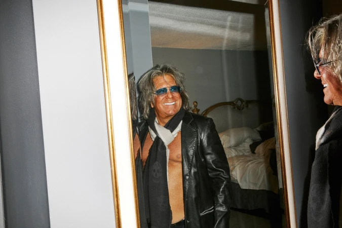Man with tinted glasses wearing a leather jacket and scarf looking into a large ornate mirror, photographed by Jeremy Liebman.