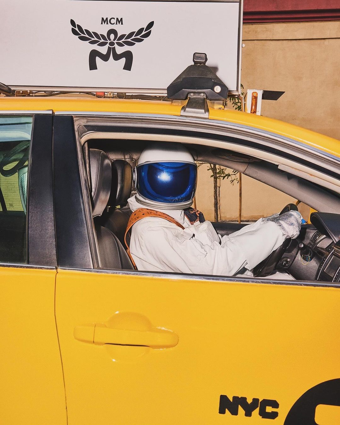 Astronaut in taxi cab with MCM signage, New York City. Yellow taxi, white space suit and helmet with dark blue visor.