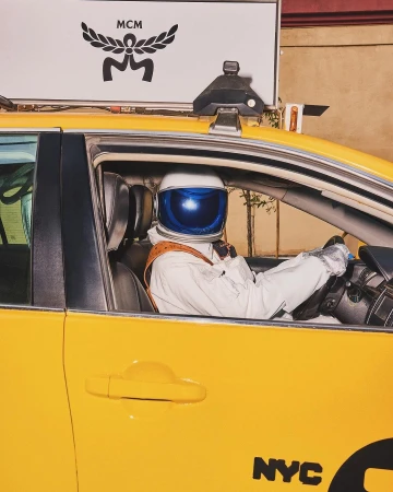 Astronaut in taxi cab with MCM signage, New York City. Yellow taxi, white space suit and helmet with dark blue visor.