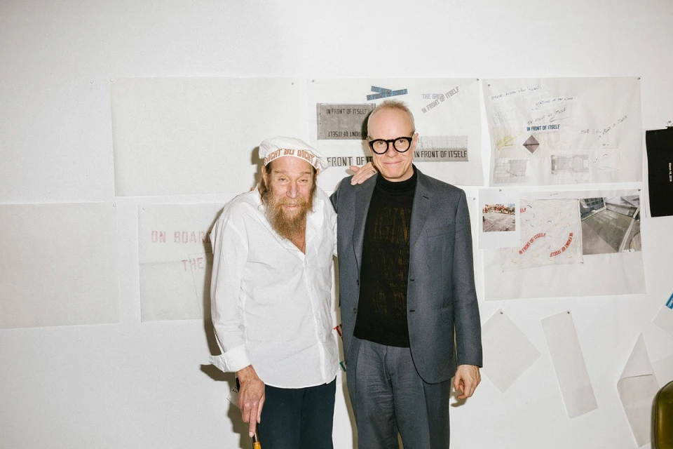 Two men pose indoors against a wall covered in whiteboards and papers with writing and diagrams, offering a glimpse into a creative studio.