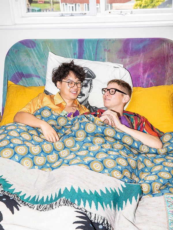 Two people wearing eyeglasses under a patterned blanket in bed with yellow pillows and a patterned headboard, interior shot.