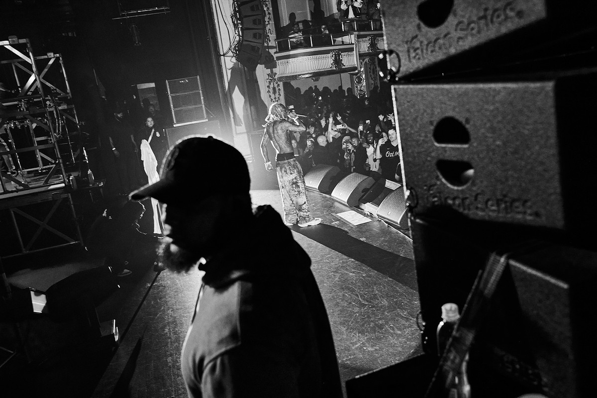 Monochromatic concert photo features a male rapper performing on stage in front of a crowd, with a hooded figure in the foreground.