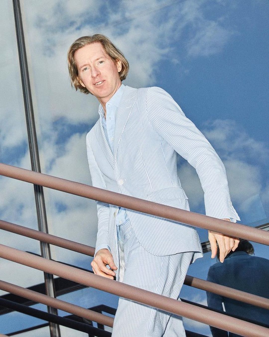 Man in a blue and white striped suit ascends outdoor stairs in daylight against a sky with fluffy white clouds, creating an elevated and stylish moment.