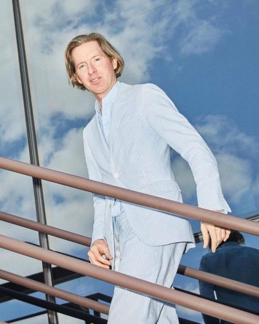 Man in a blue and white striped suit ascends outdoor stairs in daylight against a sky with fluffy white clouds, creating an elevated and stylish moment.