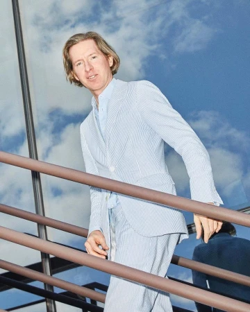 Man in a blue and white striped suit ascends outdoor stairs in daylight against a sky with fluffy white clouds, creating an elevated and stylish moment.