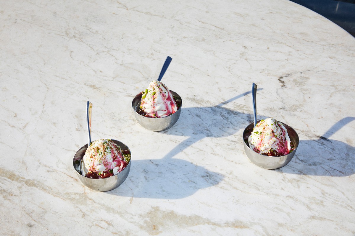 Three bowls of ice cream with syrup and pistachios sit on a marble table, each with a spoon, under bright, natural lighting.