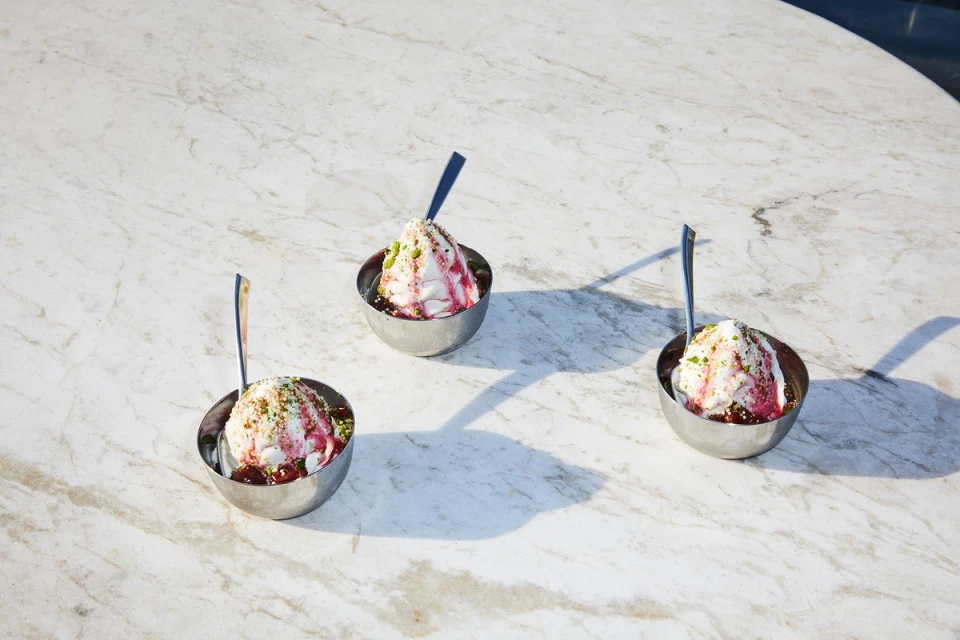 Three bowls of ice cream with syrup and pistachios sit on a marble table, each with a spoon, under bright, natural lighting.
