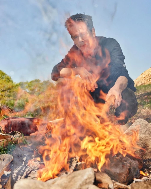 Man crouching by a fire in an outdoor setting, with rocks and greenery visible in the background under a partly cloudy sky.