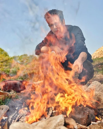Man crouching by a fire in an outdoor setting, with rocks and greenery visible in the background under a partly cloudy sky.