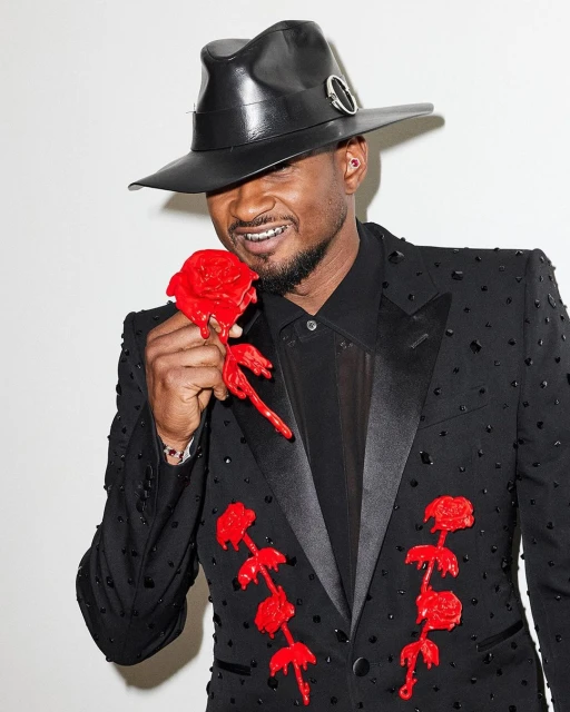 Man wearing black hat and studded suit holds a red rose. He smiles against a white background in a studio portrait.