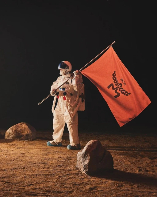 An astronaut in a white spacesuit plants an orange flag with a dark MCM logo on a sepia-toned planet-like surface with rocks.