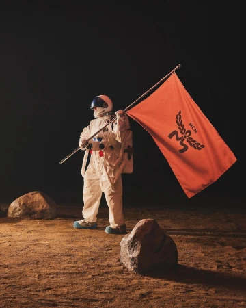An astronaut in a white spacesuit plants an orange flag with a dark MCM logo on a sepia-toned planet-like surface with rocks.