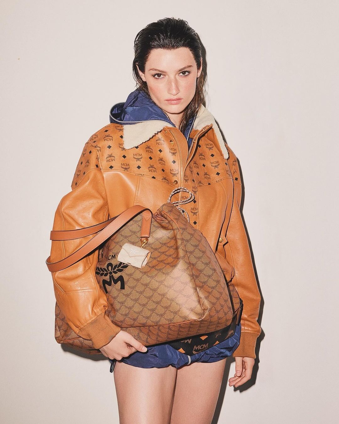 A woman in an MCM jacket and shorts holds a large MCM bag against a neutral backdrop in a fashion studio shot.