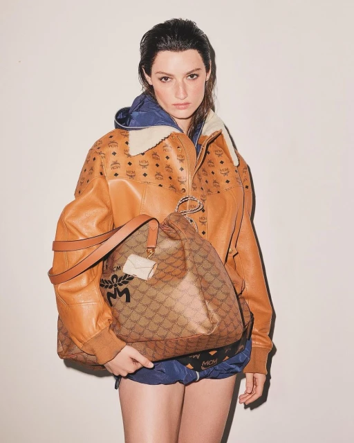 A woman in an MCM jacket and shorts holds a large MCM bag against a neutral backdrop in a fashion studio shot.