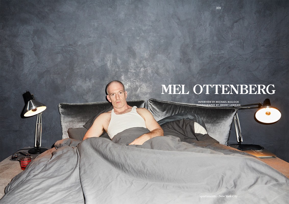 An adult man in a white tank top sits in a grey bed with black lamps on either side, against a textured grey wall in a home interior.