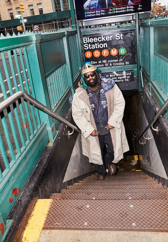 Man in trench coat standing on subway stairs in New York City. Subway sign reads Bleecker St Station, B, D, F, M, 6 lines.