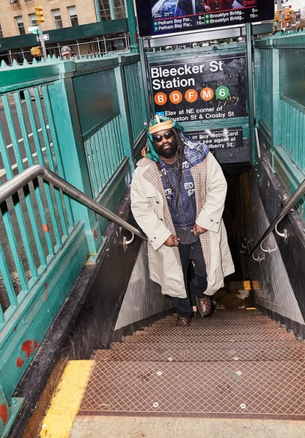 Man in trench coat standing on subway stairs in New York City. Subway sign reads Bleecker St Station, B, D, F, M, 6 lines.