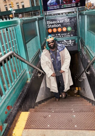 Man in trench coat standing on subway stairs in New York City. Subway sign reads Bleecker St Station, B, D, F, M, 6 lines.