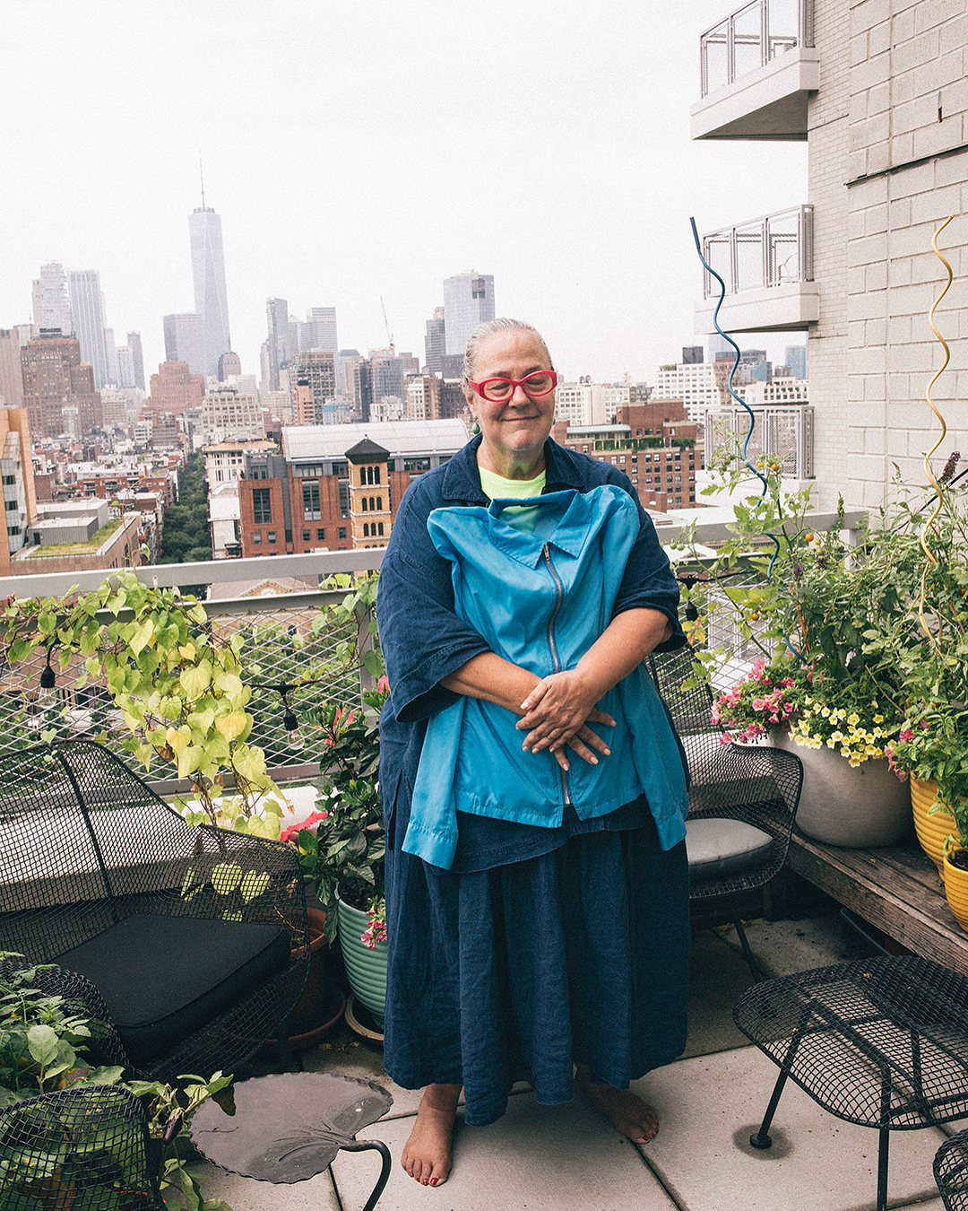 Adult woman on a balcony in a blue jacket, city skyline in the background, an outdoor scene with plants and seating.