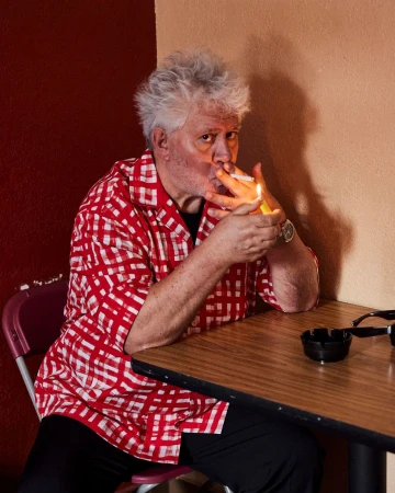 Older man with white hair in red patterned shirt sits at table lighting a cigarette with a lighter. Interior shot, indoors.