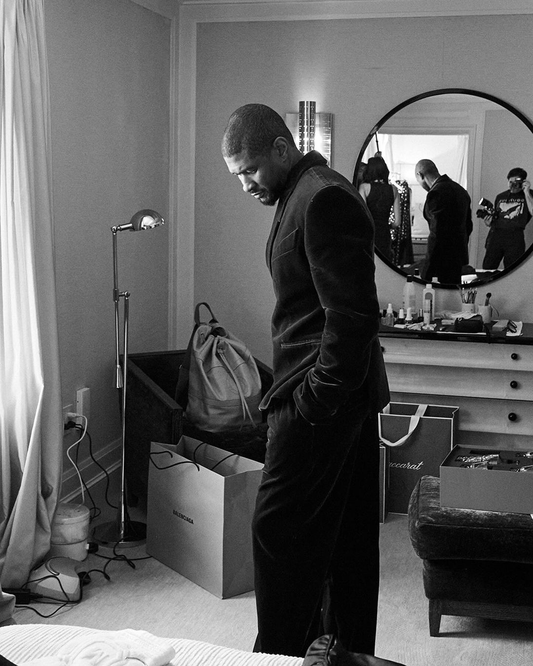 Black and white portrait of a man in a suit jacket in a hotel room with shopping bags and a dresser visible in background.