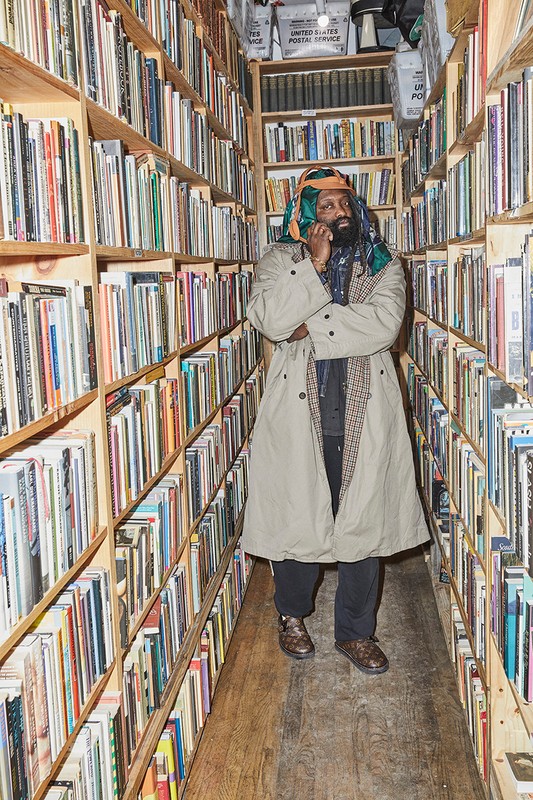 Man stands in a bookstore aisle surrounded by tall wooden shelves filled with books. He is wearing a tan coat and patterned scarf.