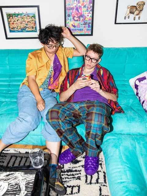 Two people pose on a teal couch in a home setting. One person uses a phone, while the other touches their hair. Framed art on the wall.