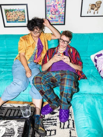 Two people pose on a teal couch in a home setting. One person uses a phone, while the other touches their hair. Framed art on the wall.