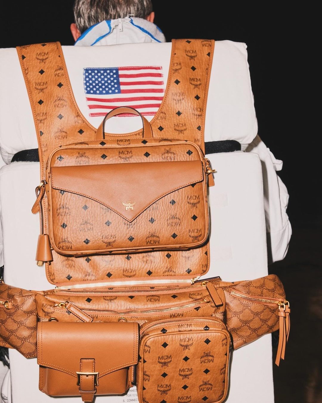 Brown MCM patterned leather backpack and waist bag with a visible American flag patch, worn with a white jacket against a dark background.