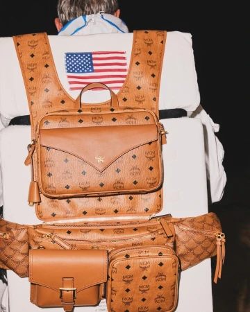 Brown MCM patterned leather backpack and waist bag with a visible American flag patch, worn with a white jacket against a dark background.